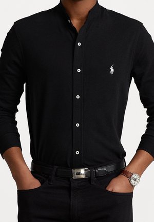 Mand iført sort skjorte med knapper med hvidt polo spiller-logo, sort bælte, sorte bukser og sølvur med brun læderrem.