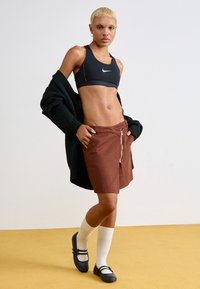 soutien desportivo preto, calções castanhos com zíper frontal, cardigan preto, meias brancas até ao joelho e sapatos pretos com fivelas num fundo amarelo.