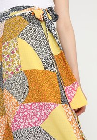 Gonna maxi patchwork nei toni del giallo, arancione, rosa e nero, con vari motivi floreali e di foglie, dotata di una cintura con dettagli a fiocco.