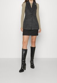 Gilet grigio su misura con un solo bottone, indossato su un top lungo a maniche ribattute color verde oliva, abbinato a una mini gonna nera e stivali neri sopra il ginocchio.