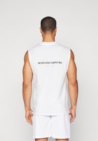 Biela bezrukávová bavlnená košeľa s okrúhlym výstrihom. Na zadnej strane sa nachádza text "NEVER STOP COMPETING" v sivej farbe, s "EST 2017" pod ním.