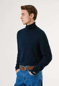 Jūrinės turtleneck megztinis, pagamintas iš minkštos medžiagos, su ilgomis rankovėmis ir raukšlėmis. Pora su mėlynomis džinsomis ir rudos odos diržu.