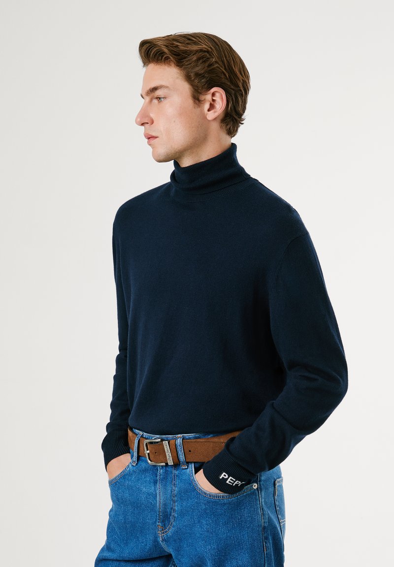 Granatowy sweter w serek wykonany z miękkiego materiału, z długimi rękawami i prążkowanymi mankietami. Połączony z niebieskimi dżinsami i brązowym skórzanym paskiem.