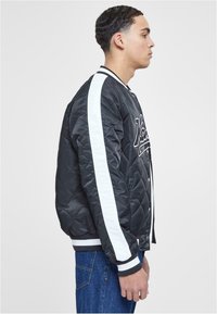 Black quilted bomber jacket with white stripes on sleeves. Features a zip closure and ribbed cuffs and hem. Accented with embroidered text.