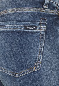 Pantalones vaqueros de mezclilla con lavado oscuro, que cuentan con un pequeño bolsillo trasero. Costuras naranjas a lo largo de las costuras. La etiqueta dice “Marc O'Polo DENIM.”
