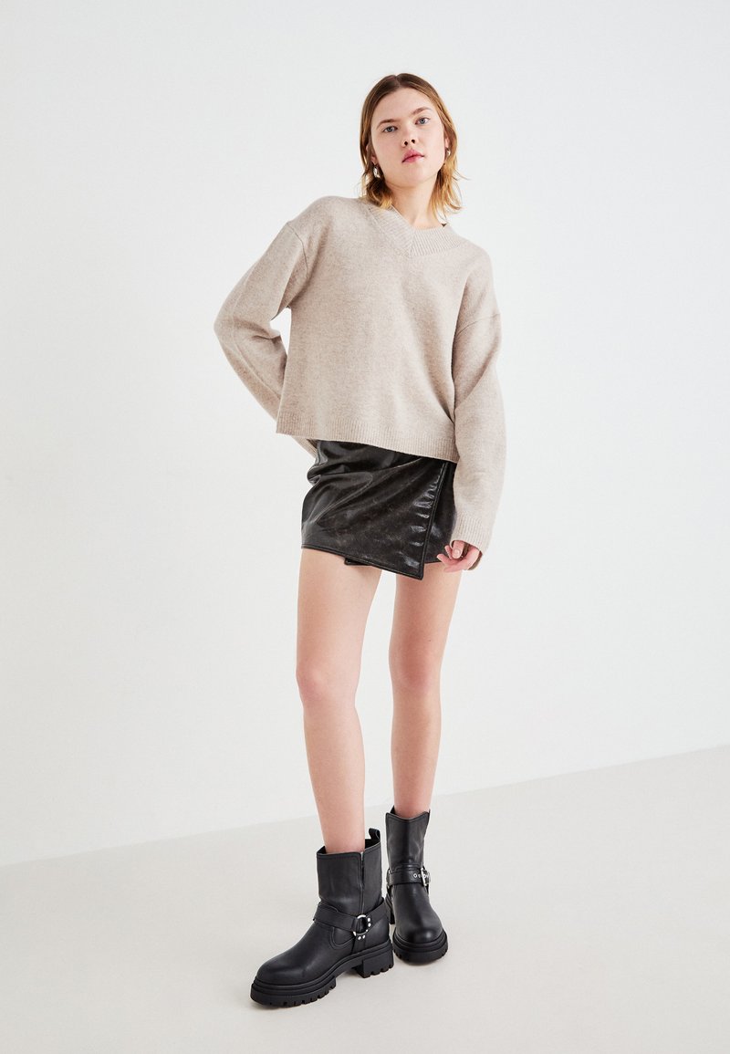 Młoda kobieta stoi w beżowym swetrze, czarnej skórzanej mini spódnicy i czarnych botkach za kostkę na jednolitym białym tle.