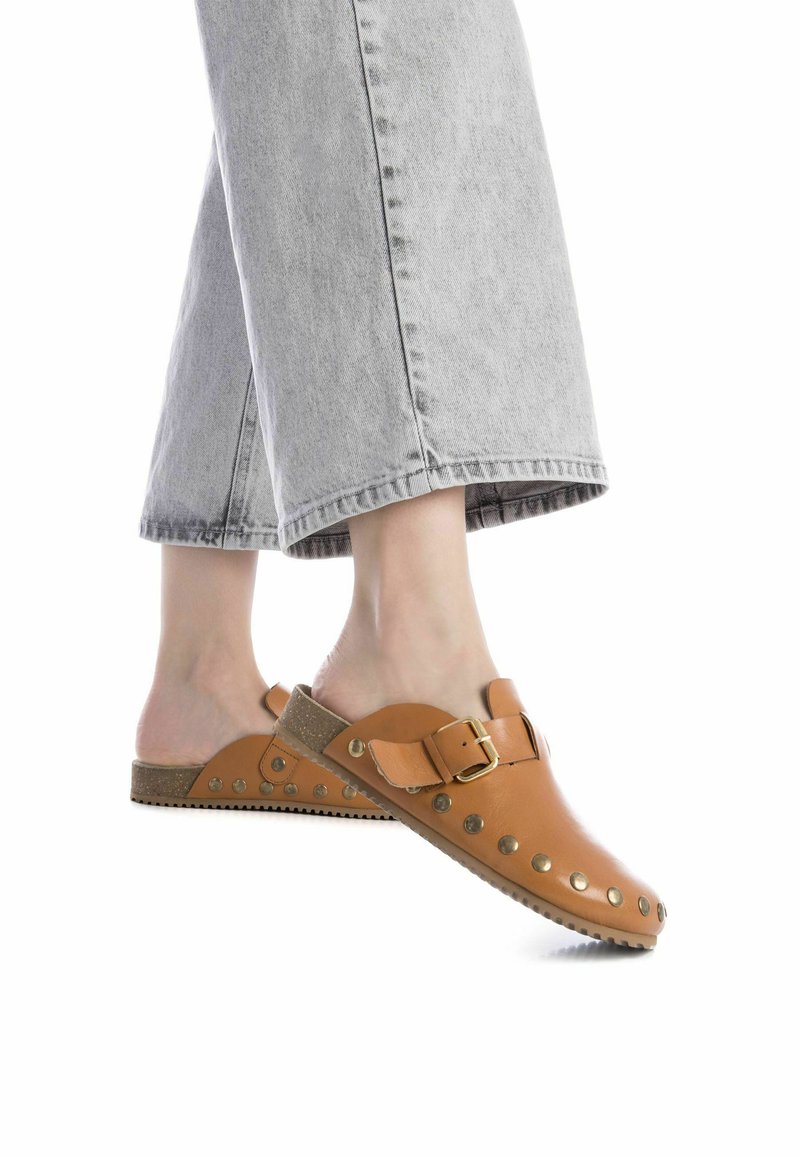 Zuecos de cuero marrón con un hebilla lateral, suela texturizada y tachuelas metálicas. Llevados con pantalones de pierna ancha gris claro, mostrando un diseño minimalista.