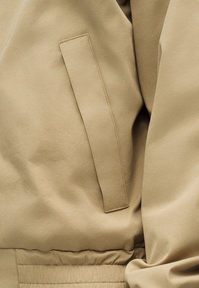 Κοντινή λήψη υφάσματος μπεζ σακακιού που δείχνει μια κάθετη τσέπη welt με ορατή ραφή και ελαστική μέση που μαζεύει από κάτω.