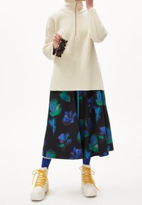 Pull col roulé en crème côtelé, jupe noire avec des motifs floraux en bleu et vert, chaussettes bleues et bottines blanches à lacets avec des accents jaunes.