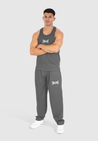 Grå tanktop og matchende sweatpants lavet af blødt stof med et hvidt logo. Modellen står med armene korslagte og bærer hvide sneakers.