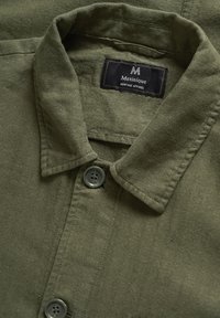Olīvzaļš krekls ar apkakli, kam ir zāģveida tekstūra, divi pelēki pogas un etiķete ar uzrakstu "Matinique Heritage Apparel".