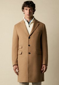 Kamelhaar-Mantel mit maßgeschneiderter Silhouette, gekerbten Revers, drei vorderen Knöpfen und zwei aufgesetzten Taschen. Getragen über einer cremefarbenen Strickschicht.