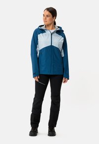 Giacca a vento blu e azzurro chiaro con cappuccio, dotata di tasche con zip e un design a pannelli. Abbinata a pantaloni neri e stivali robusti.