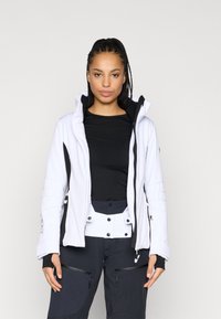 Λευκή σακάκι σκι με μαύρες λεπτομέρειες, φερμουάρ μπροστά, ψηλό γιακά και υφασμάτινα μανίκια. Συνδυασμένο με σκούρα παντελόνια με τσέπες με φερμουάρ.