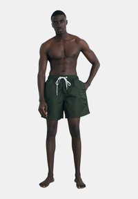 Costumi da bagno verdi realizzati in tessuto leggero, con vita regolabile da cordino con dettagli dorati, lunghezza sopra il ginocchio e texture liscia.