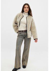 Beige fleece-takki mustalla vetoketjulla ja valkoisilla yksityiskohdilla, yhdistettynä kermaiseen poolopaitaan ja harmaisiin leveälahkeisiin farkkuihin. Mustat nilkkurit viimeistelevät asun.
