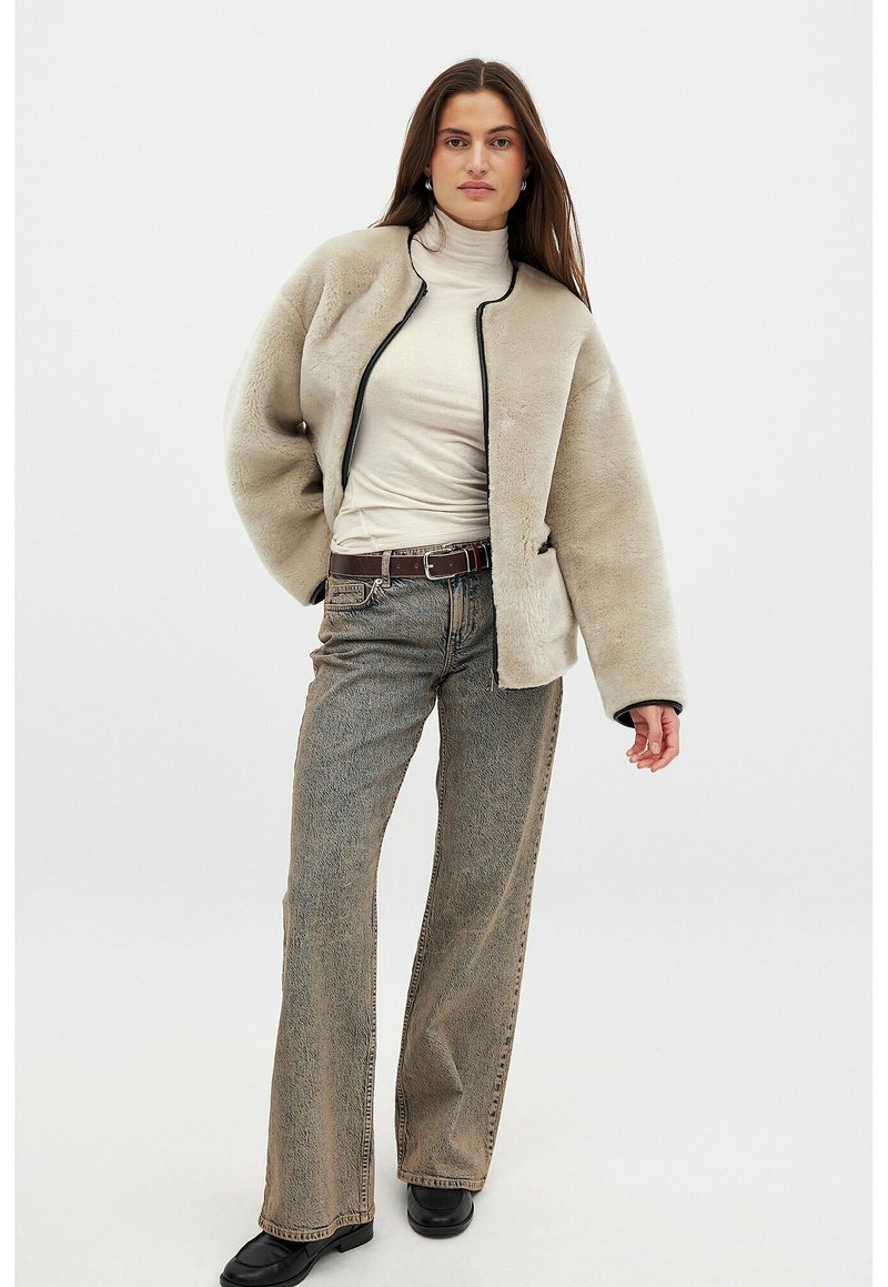 Beige fleece-takki mustalla vetoketjulla ja valkoisilla yksityiskohdilla, yhdistettynä kermaiseen poolopaitaan ja harmaisiin leveälahkeisiin farkkuihin. Mustat nilkkurit viimeistelevät asun.