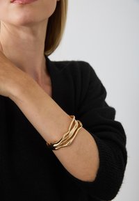 Bracelet en métal doré avec un design ondulé et entrelacé. Fini lisse et surface réfléchissante, porté sur un poignet nu avec un pull noir.