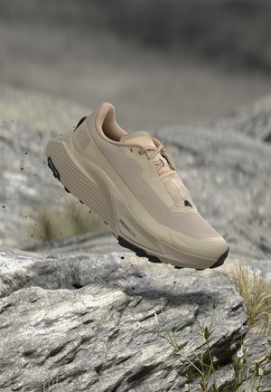 Chaussure de randonnée beige avec semelle noire en l'air au-dessus d'un terrain rocheux avec de l'herbe et de petites fleurs blanches en arrière-plan.