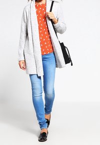 Grijze gebreide cardigan, oranje bloes met bloemen, blauwe skinny jeans en zwarte instappers. De outfit heeft een casual gelaagde uitstraling.