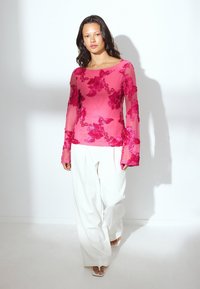 Blusa rosa chiaro con maniche lunghe a campana in tessuto trasparente e motivi floreali ricamati in tonalità vivaci di rosa. Abbinata a pantaloni bianchi a gamba larga.
