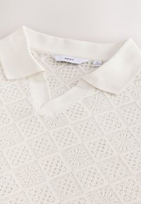 Pull-over blanc en maille avec un col en V, présentant un motif en losange texturé et un col côtelé doux et contrastant. Étiquette de marque visible à l'intérieur.