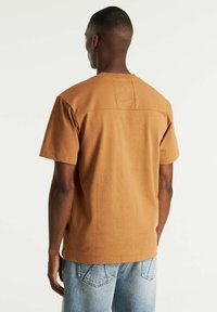 Brun bomulds t-shirt med korte ærmer og rund halsudskæring, med en lille accentsting på ryggen. Sammen med lyseblå jeans.