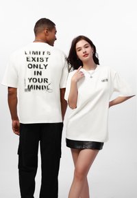 Weiße Baumwoll-T-Shirts, die auf zwei Modellen präsentiert werden, verfügen über auffällige Textdrucke auf der Vorder- und Rückseite, mit lockerer Passform und kurzen Ärmeln.