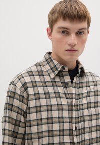 Una camisa de cuadros beige y verde hecha de tela suave, con cierre de botones en la parte frontal, cuello y un bolsillo de parche en el lado izquierdo.