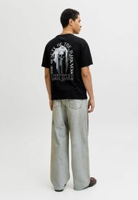 Czarny t-shirt z grafiką czaszki i napisem na plecach. Połączony z jasnoszarymi szerokimi spodniami dżinsowymi. Model pokazany z tyłu.
