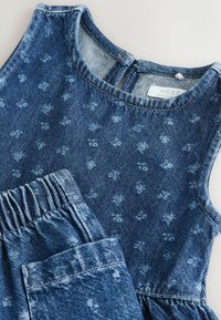 Jeans obleka v temno modri barvi s cvetličnim vzorcem, ki vključuje brezrokavnik in hlače z elastičnim pasom. Mehka tekstura in sproščen dizajn.