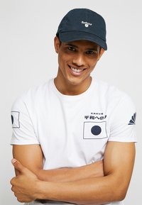 Marineblauwe baseballpet met "Barbour" logo, witte T-shirt met grafische prints waaronder een vlagontwerp, model met gekruiste armen, lichte achtergrond.