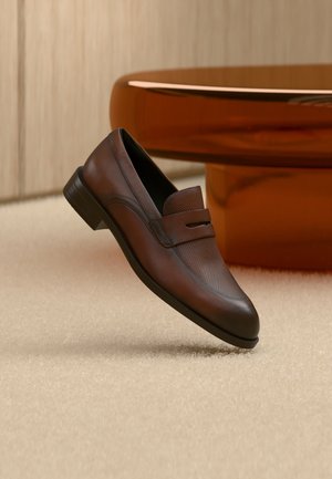 Pruun nahast meeste loafer, mille eesosas on augukesed ja rihma detail, asetatud beeži vaiba kohale, taustal ümmargune läikiv pruun laud.