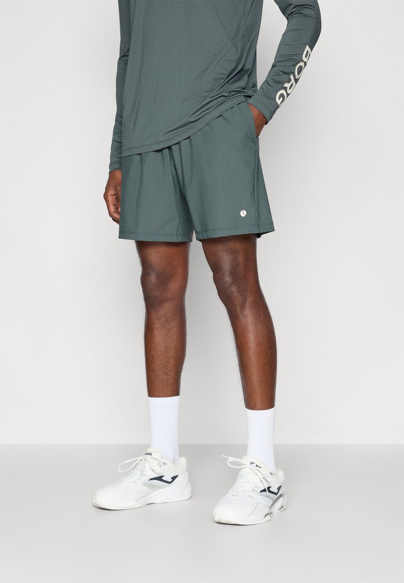 Σκούρο πράσινο αθλητικό σορτς, ελαφρύ υλικό, ελαστική μέση, πλαϊνές τσέπες, λεπτομέρεια λογότυπου; συνδυασμένο με λευκά αθλητικά παπούτσια και κάλτσες.