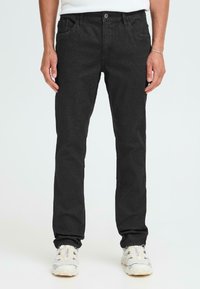 Črne jeans hlače z ožjim krojem, petimi žepi in gumbom za zapiranje. Teksturovan material z diskretnimi šivi; oblečene z belimi supergami.