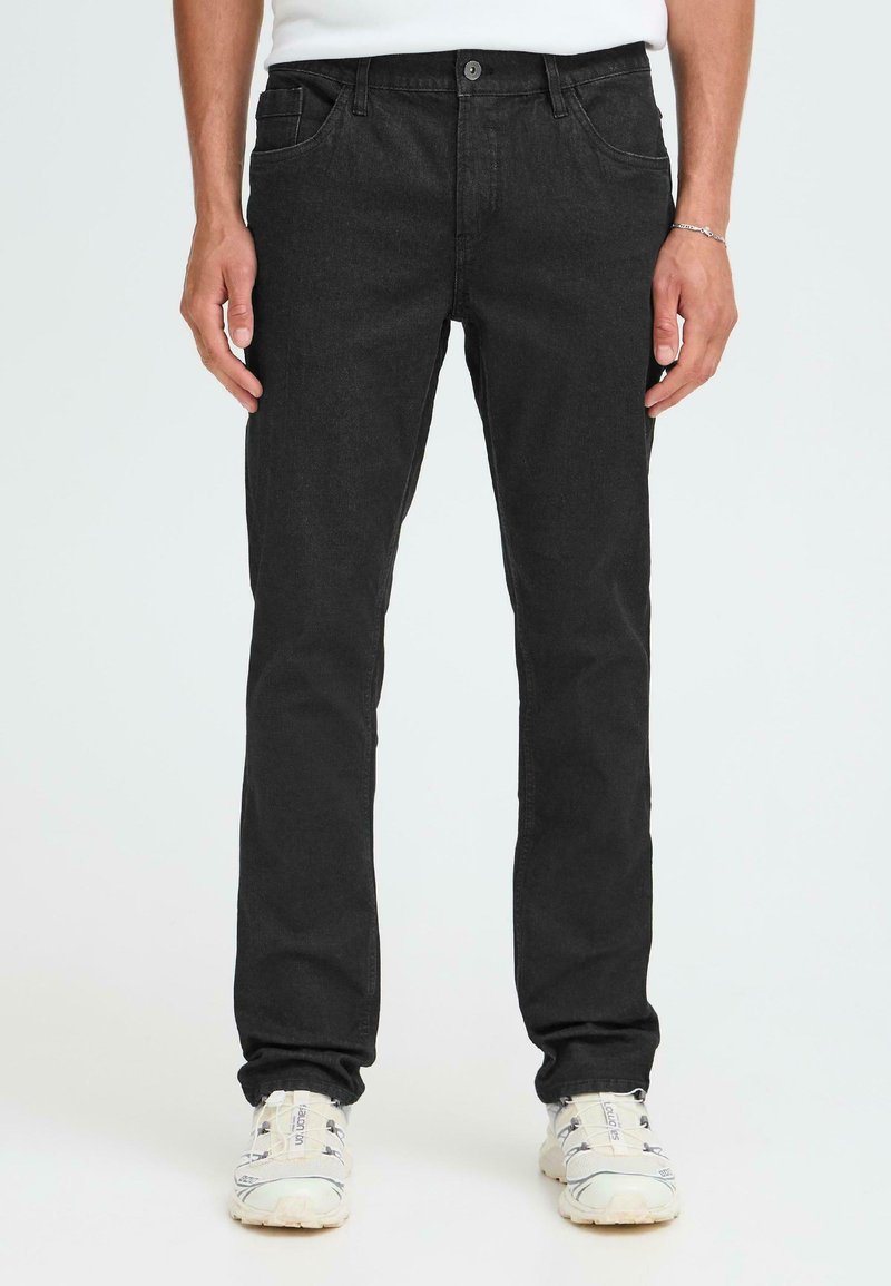 Črne jeans hlače z ožjim krojem, petimi žepi in gumbom za zapiranje. Teksturovan material z diskretnimi šivi; oblečene z belimi supergami.