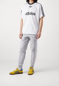 Vit kortärmad Adidas-tröja med svart logotyp, tillsammans med grå joggingbyxor med vita ränder, och gula sneakers med blå detaljer.