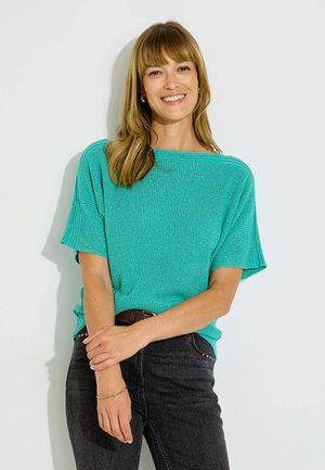 Femme aux cheveux châtain clair mi-longs sourit en portant un haut tricoté turquoise à manches courtes et un jean noir avec une ceinture marron.
