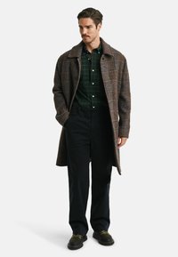 Cappotto a quadri marrone sopra una camicia a quadri verde, pantaloni neri e scarpe scure. Il cappotto è lungo fino al ginocchio, con un grande colletto e tasche laterali.