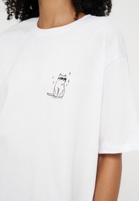 T-shirt blanc avec un petit dessin au trait noir d'un chat portant des lunettes de soleil et des paillettes sur le haut de la poitrine.