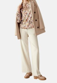 Beżowy płaszcz, kwiatowa bluzka w pastelowe wzory, kremowe szerokie spodnie oraz brązowe mokasyny z detalem łańcuszka.