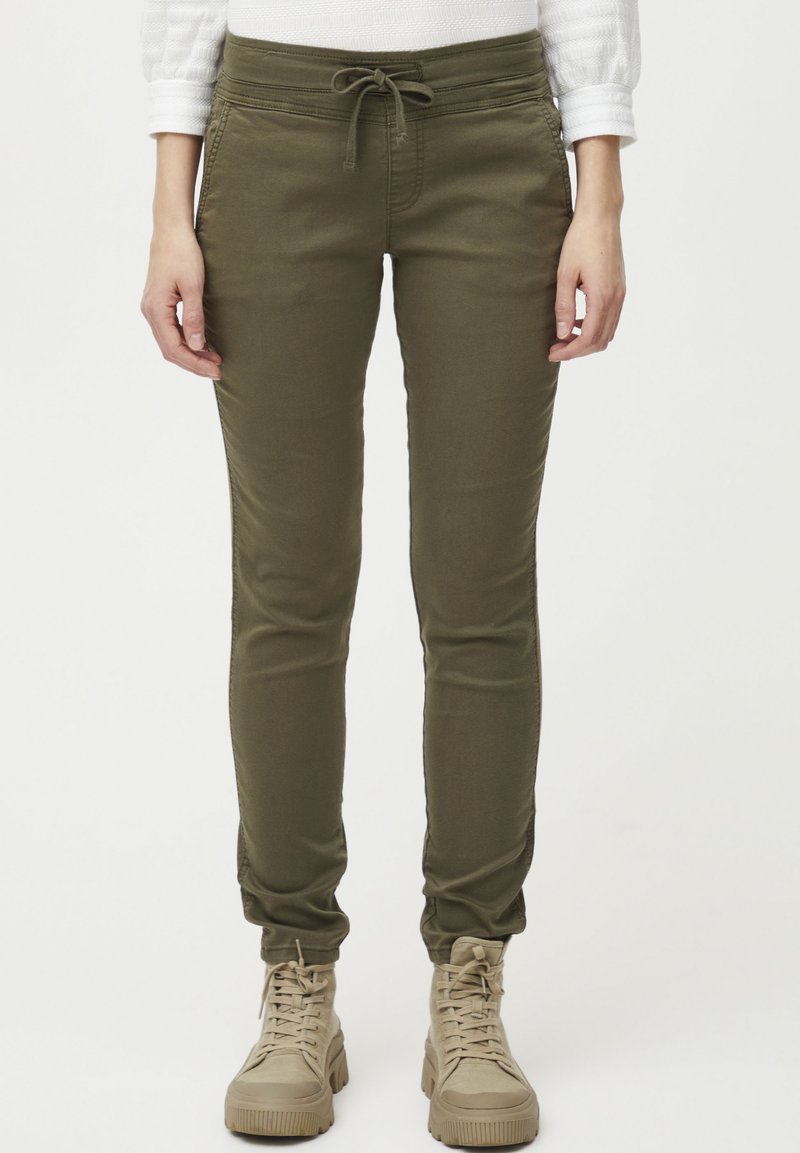 Pantalons de jogging vert olive avec une taille à cordon, jambe fuselée et poignets au niveau des chevilles. Fabriqués en tissu doux et extensible. Portés avec des baskets beiges.