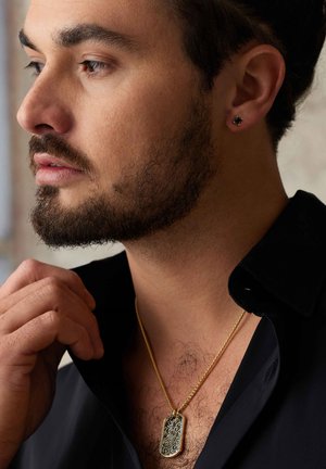 Pendentif en dog tag en or avec des gravures complexes, porté sur une chaîne en or, devant un fond de chemise noire à col.