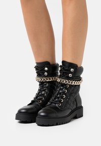 Botas de cuero negro hasta el tobillo con textura acolchada, acento de cadena dorada, parte delantera con cordones, herrajes metálicos y diseño de suela robusta.