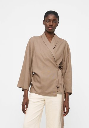 Γυναίκα που φοράει μια φαρδιά, μπεζ μπλούζα τύπου φιόγκος με φαρδιά μανίκια και λευκό παντελόνι, στέκεται μπροστά σε ένα απλό ανοικτόχρωμο φόντο.