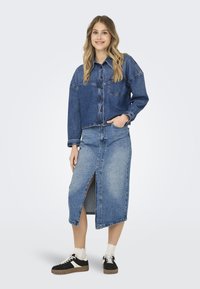 Jeans jakna s žepom, v kombinaciji z dolgo jeans krilo z sprednjim razporkom. Model nosi črne superge in bele nogavice.