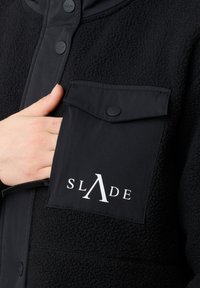 Giacca in pile nera con tasca sul petto e chiusura frontale con bottoni; la tasca ha un lembo nero con il testo bianco "SLADE".