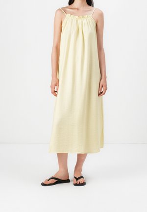 Żółta sukienka bez rękawów z marszczonym dekoltem i fakturowaną tkaniną, sięgająca do połowy łydki. W połączeniu z czarnymi klapkami.