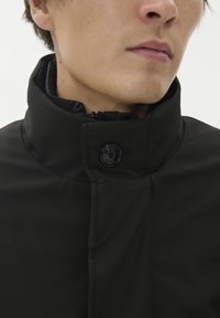 Črna jakna z visokim ovratnikom, ki ima zadrgo in gumbasto zapiranje. Gladka tekstura s subtilnimi šivi. Minimalističen dizajn.