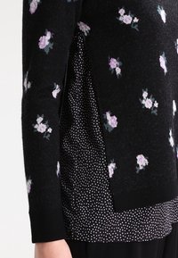 Pull noir à motif floral, superposé à une chemise noire à pois. Tricot texturé, avec de petites fentes sur les côtés. Look moderne et décontracté.